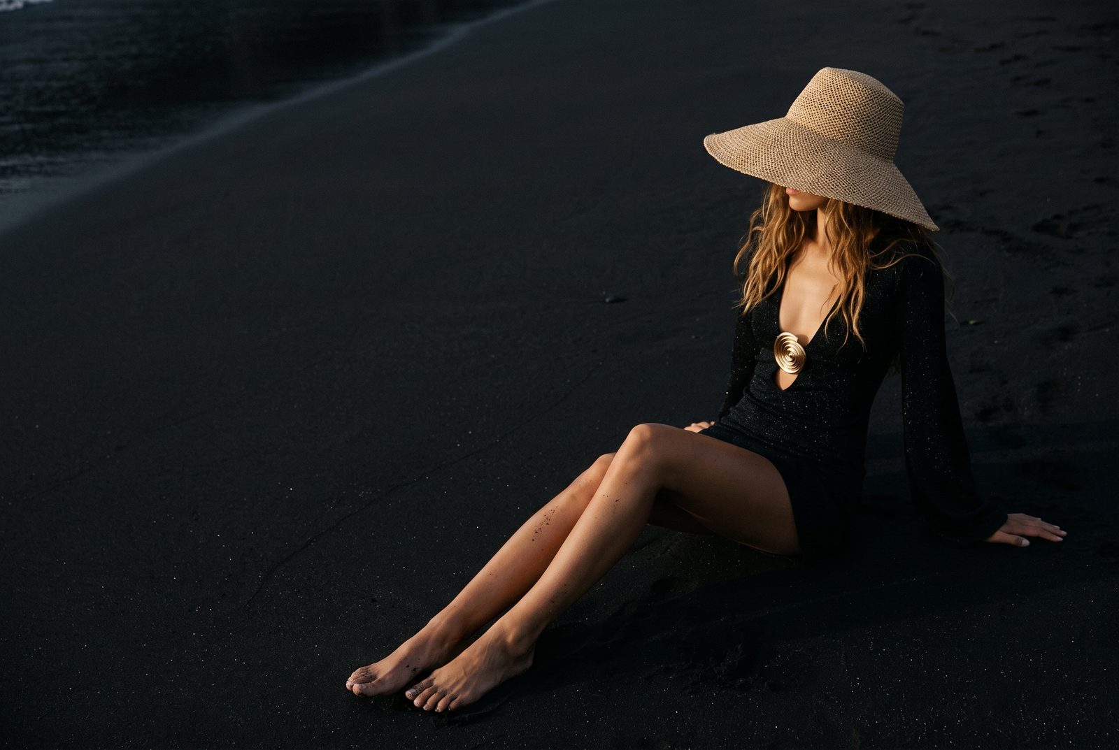 How to dress for a European summer — a woman in a straw hat seated on dark volcanic sand in the warm Mediterranean light, the ESVRA European summer edit