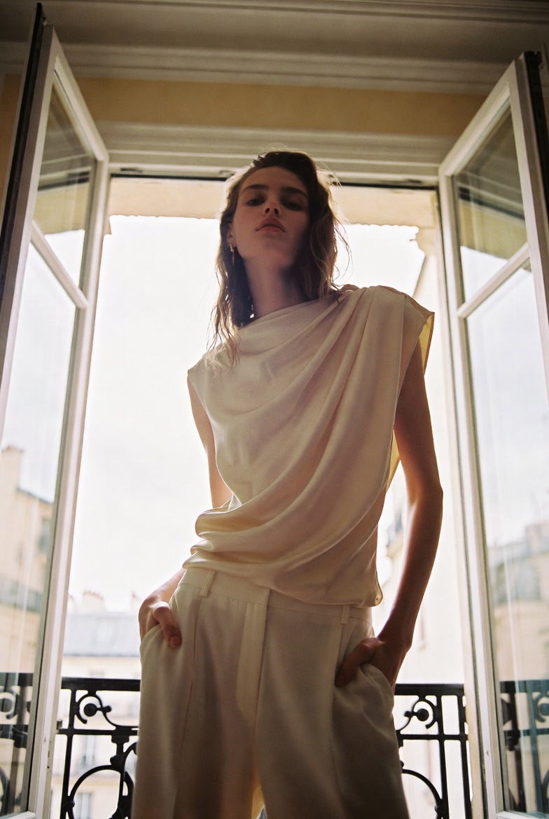 The Going-Out Top — a tall slender model in a draped silk-satin evening top and wide-leg cream trousers, leaning against a Paris apartment window, the ESVRA Styling edit on Fall 2026 evening tops