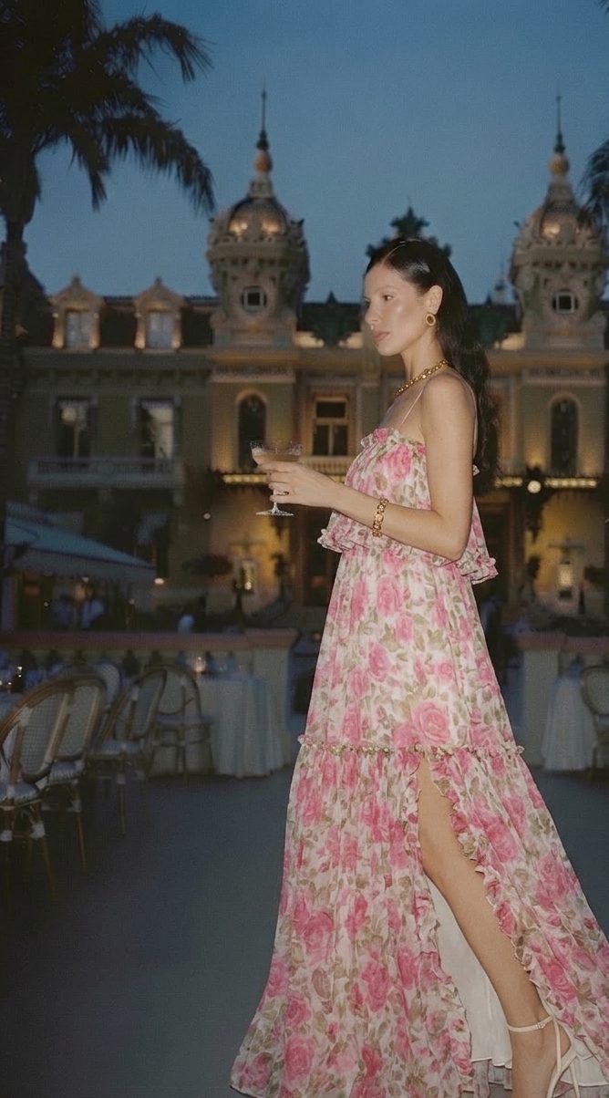 A woman in a pink rose-print chiffon maxi dress with a thigh slit holding a coupe in a Riviera square at blue-hour dusk