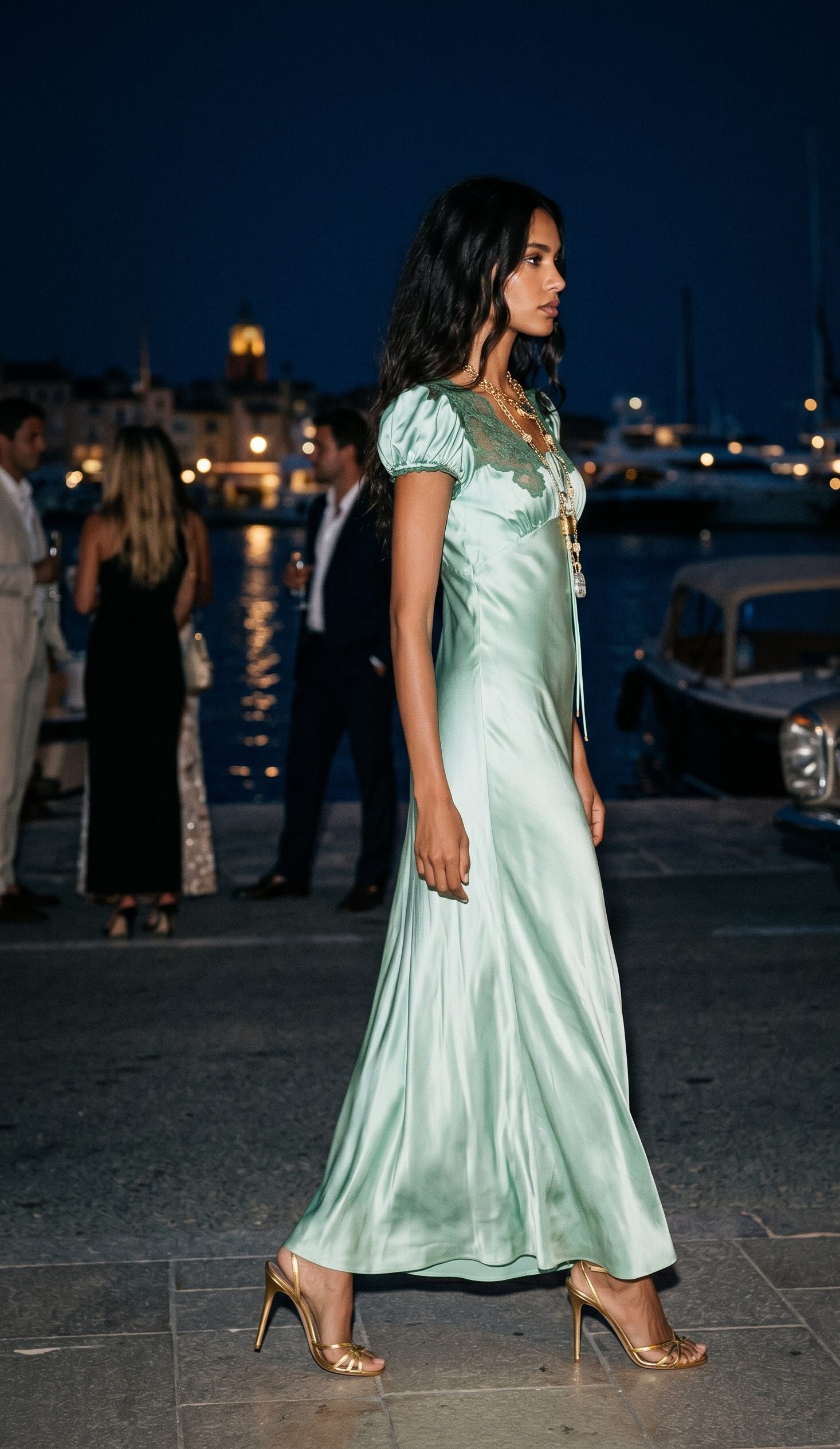 A woman in a sea-glass green bias-cut silk slip gown with gold heels walking the Saint-Tropez harbour at night past lit yachts and a vintage car
